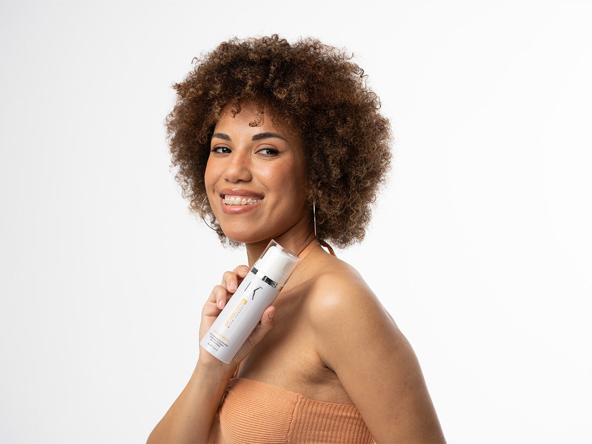 A lady with curly hair holds a leave-in conditioner, showcasing a modern beauty routine against a clean background.