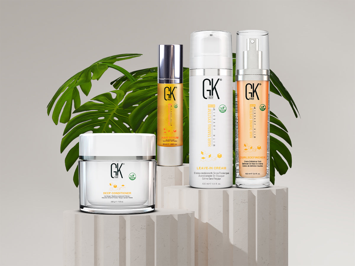 Four GK Hair products including deep conditioner, hair serum, leave-in cream, and curl definer on white pedestals with green tropical leaves backdrop.