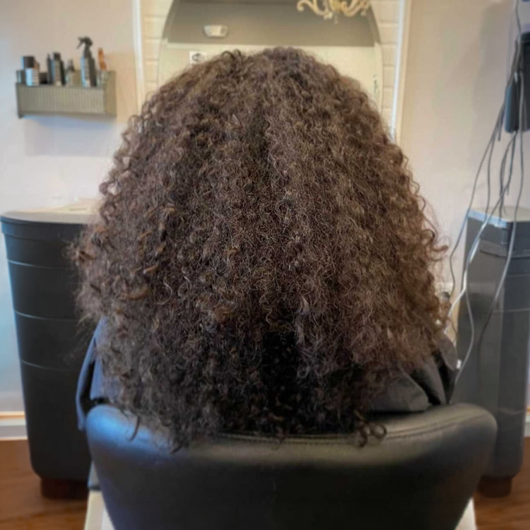 Before: tightly curly frizzy dark brown hair from behind in a salon chair, styling station and mirror behind